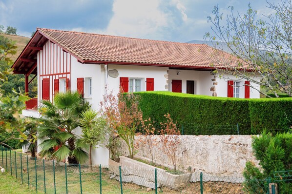 A white building with red shutters and a tiled roof, surrounded by greenery and a stone wall.