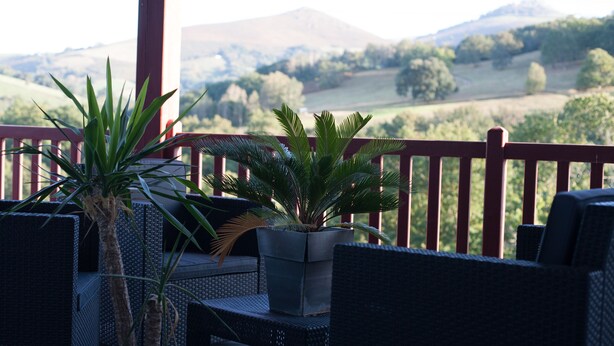 A balcony with wicker furniture and potted plants, overlooking a scenic landscape with hills and trees.