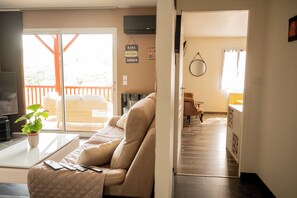 A living room with a sofa, a coffee table, and a television. There is a balcony with a view of greenery.
