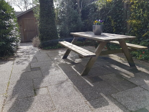 Una mesa de picnic de madera con una maceta encima, ubicada en una zona pavimentada con un cobertizo de madera y árboles al fondo.