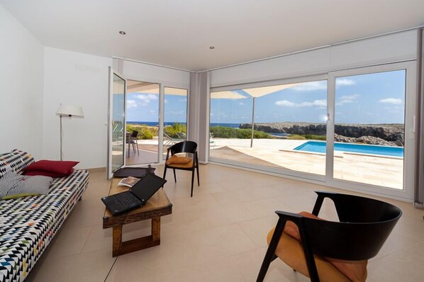 A modern living room with a sofa, a table with a laptop, and a chair. Large windows offer a view of a pool and landscape. - Panoramic Sea Views, 20m easy access to the sea, ideal for snorkelling (Es Mercadal)
