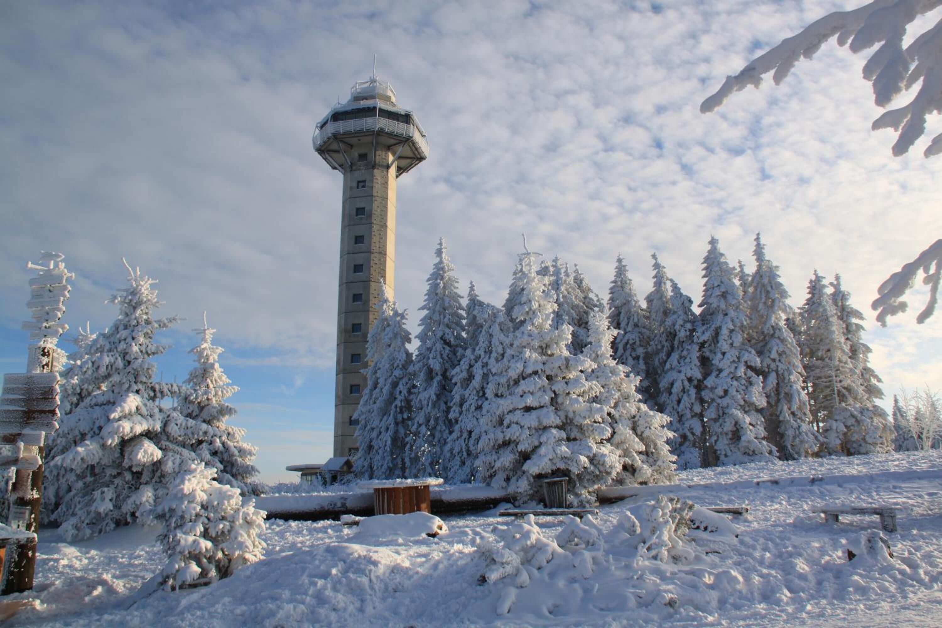 A snow-covered tower with a cylindrical shape and a rounded top, surrounded by snow-laden trees and a clear sky.