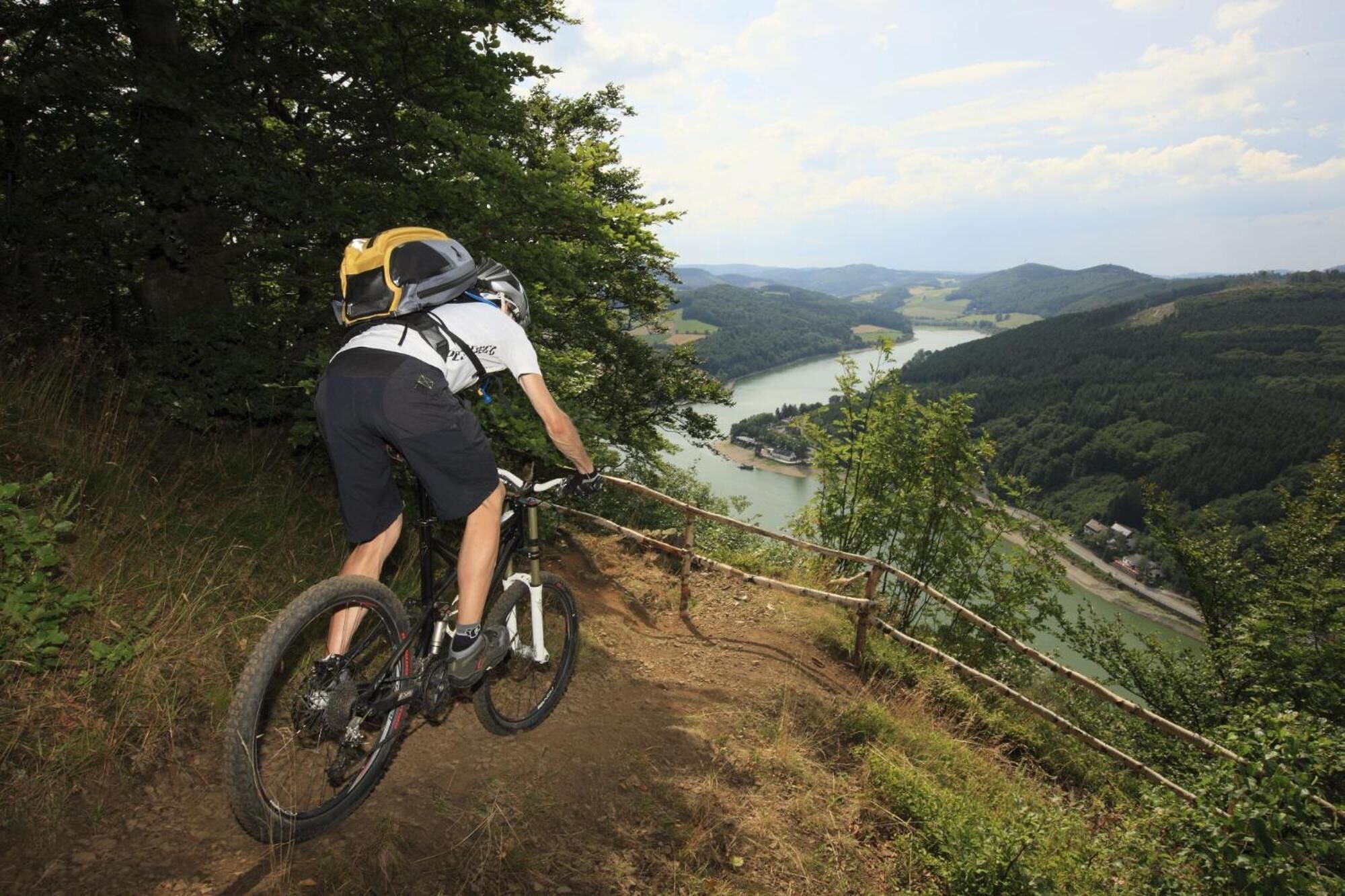 A mountain biker on a dirt trail with a scenic view of a river and hills in the background.