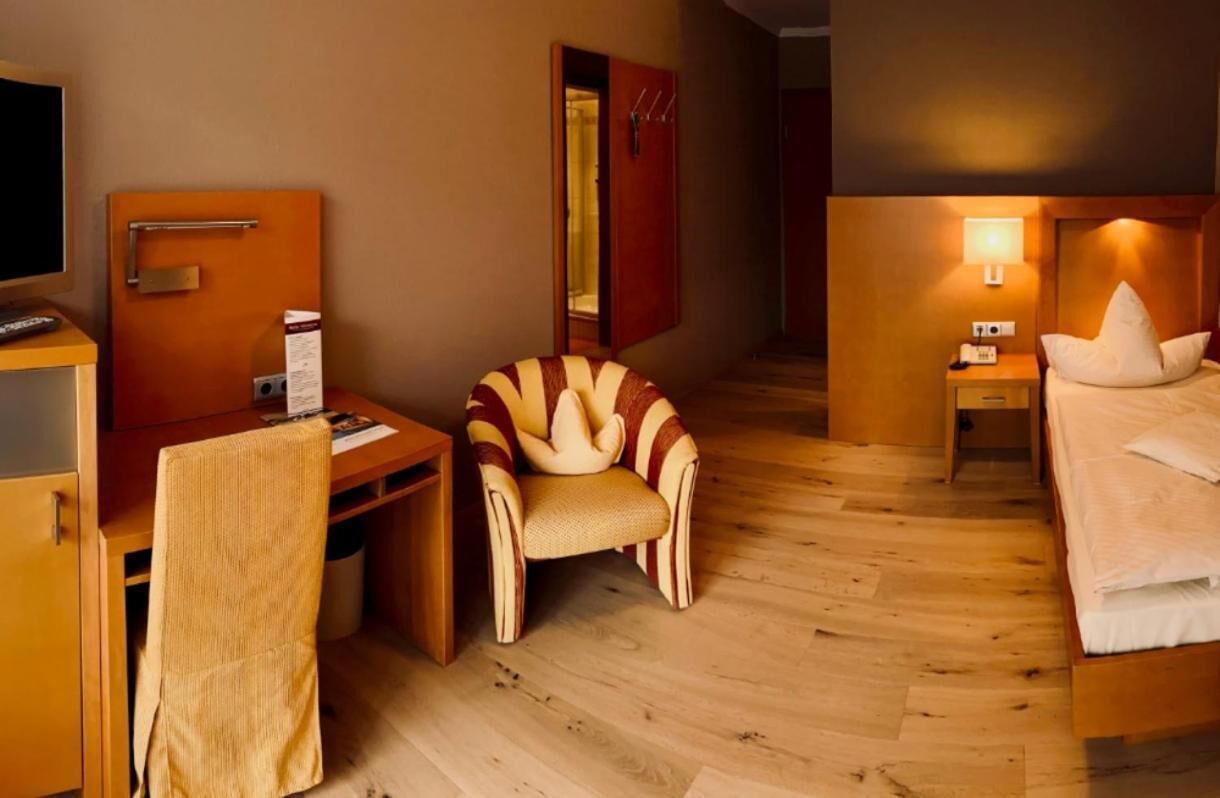 A hotel room with a wooden desk, a chair, a bed, a nightstand, and a lamp.