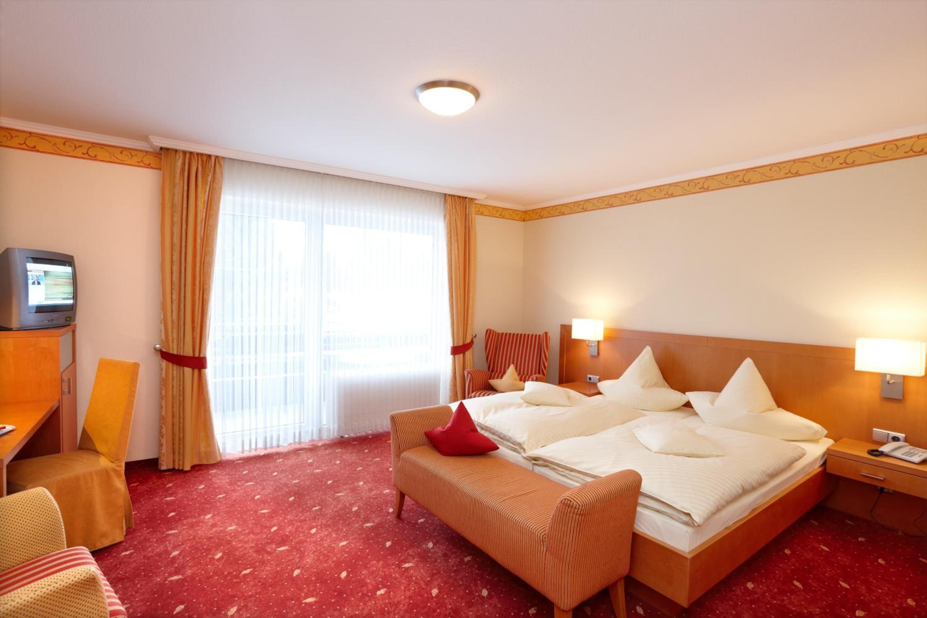 A hotel room with a large bed, a desk with a television, a chair, and a bench.