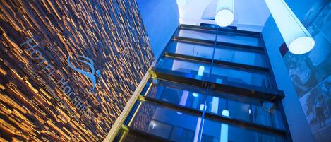 A modern hotel interior with a wooden textured wall, large windows, and hanging cylindrical lights.