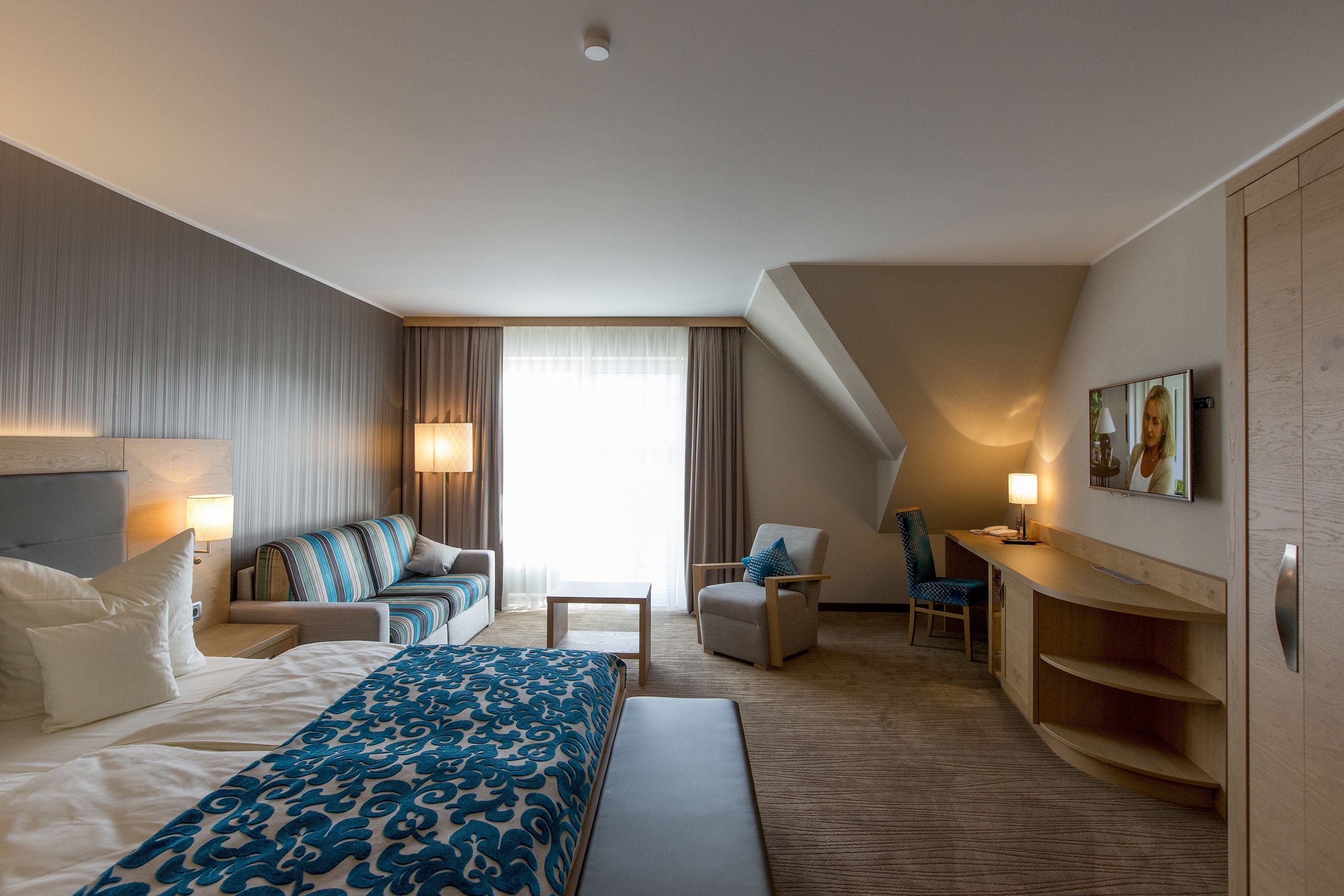 Een hotelkamer met een groot bed, een slaapbank, een bureau en een stoel. Er is een raam met gordijnen en een schilderij aan de muur.