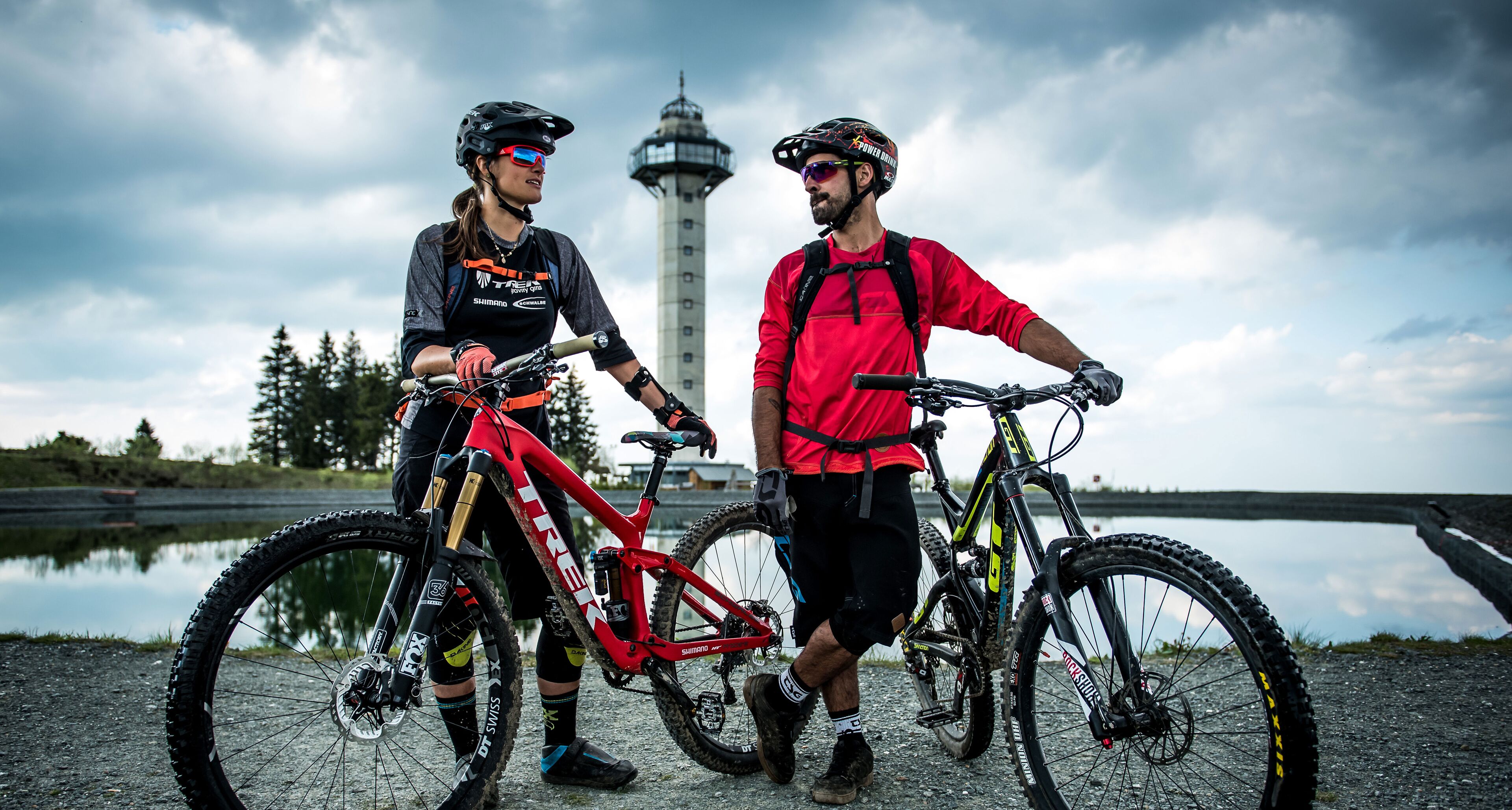 Two cyclists with mountain bikes near a body of water and a tower in the background.