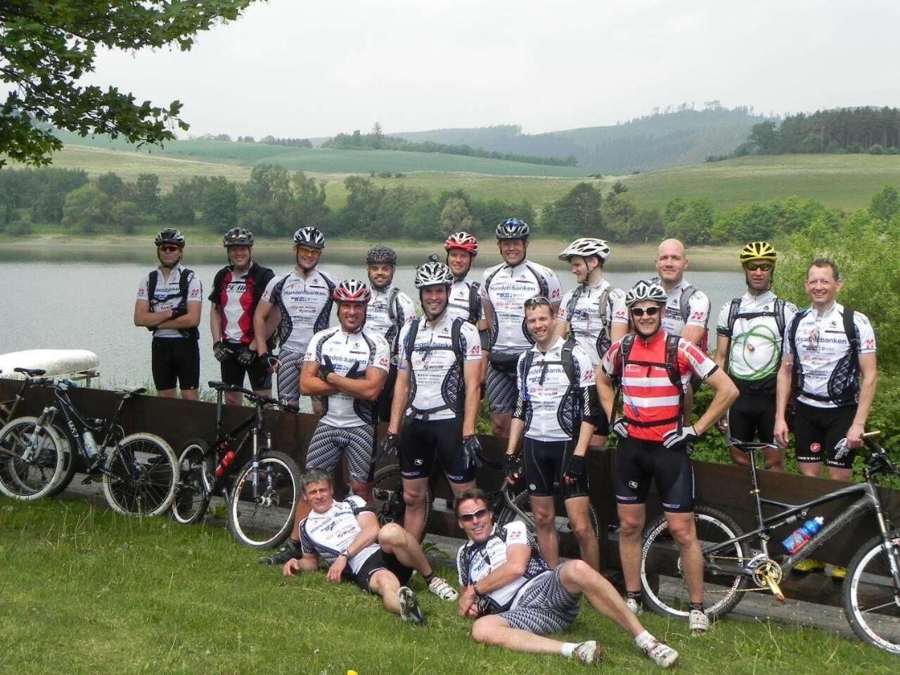 A group of cyclists posing by a lake with bicycles and a wooden fence in the background.