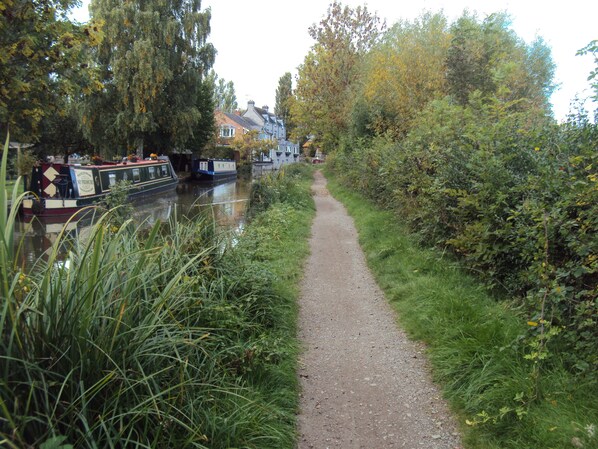 A narrow path beside a canal with boats, surrounded by greenery and trees.