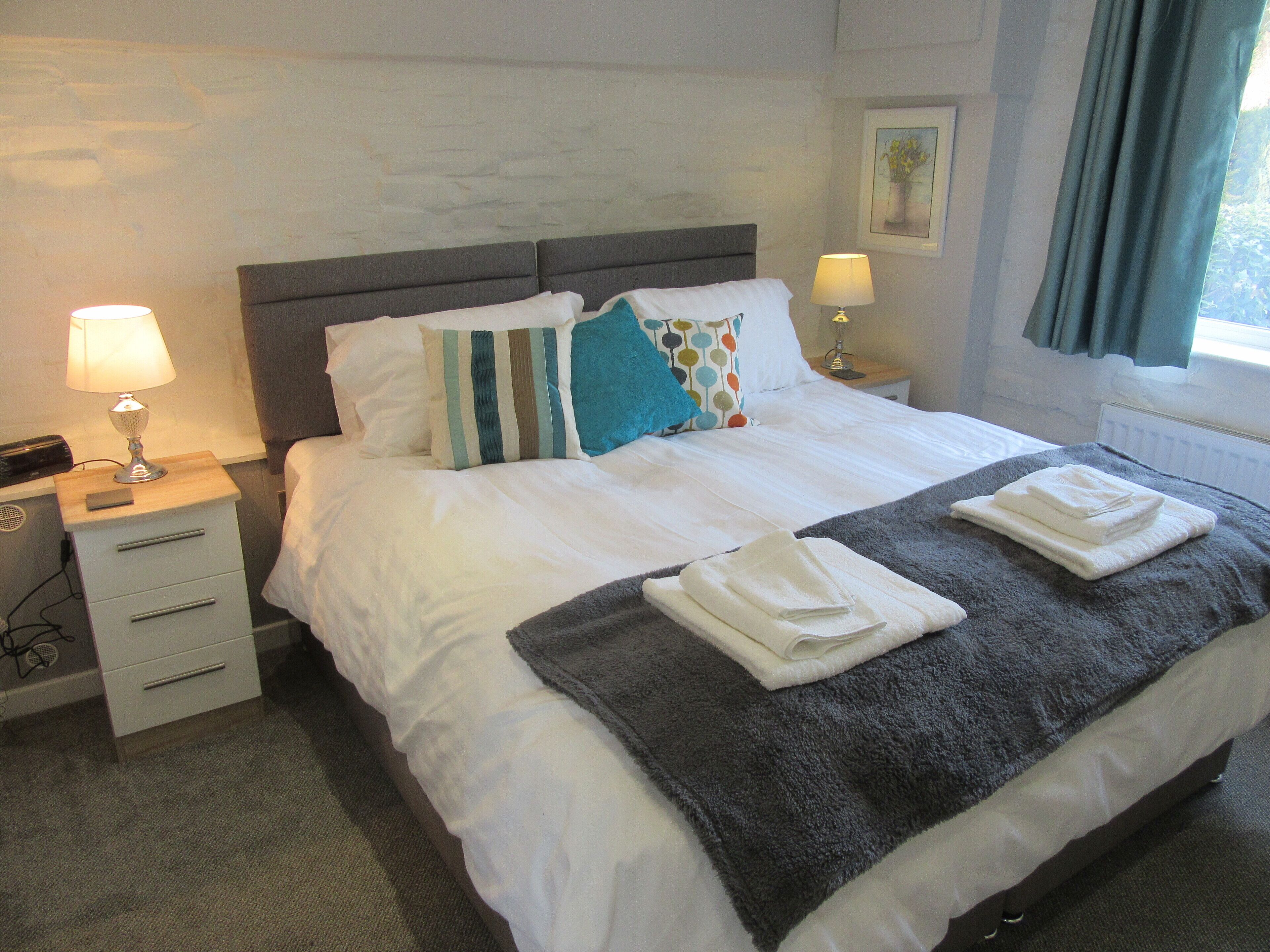 A neatly made bed with white linens, a grey throw blanket, and folded towels. There are two bedside lamps, a small bedside table, and a wall-mounted picture.