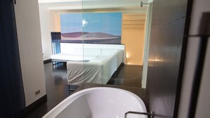 A modern bathroom with a large bathtub, a bed with a white bedspread, and a large window with a view of a landscape.