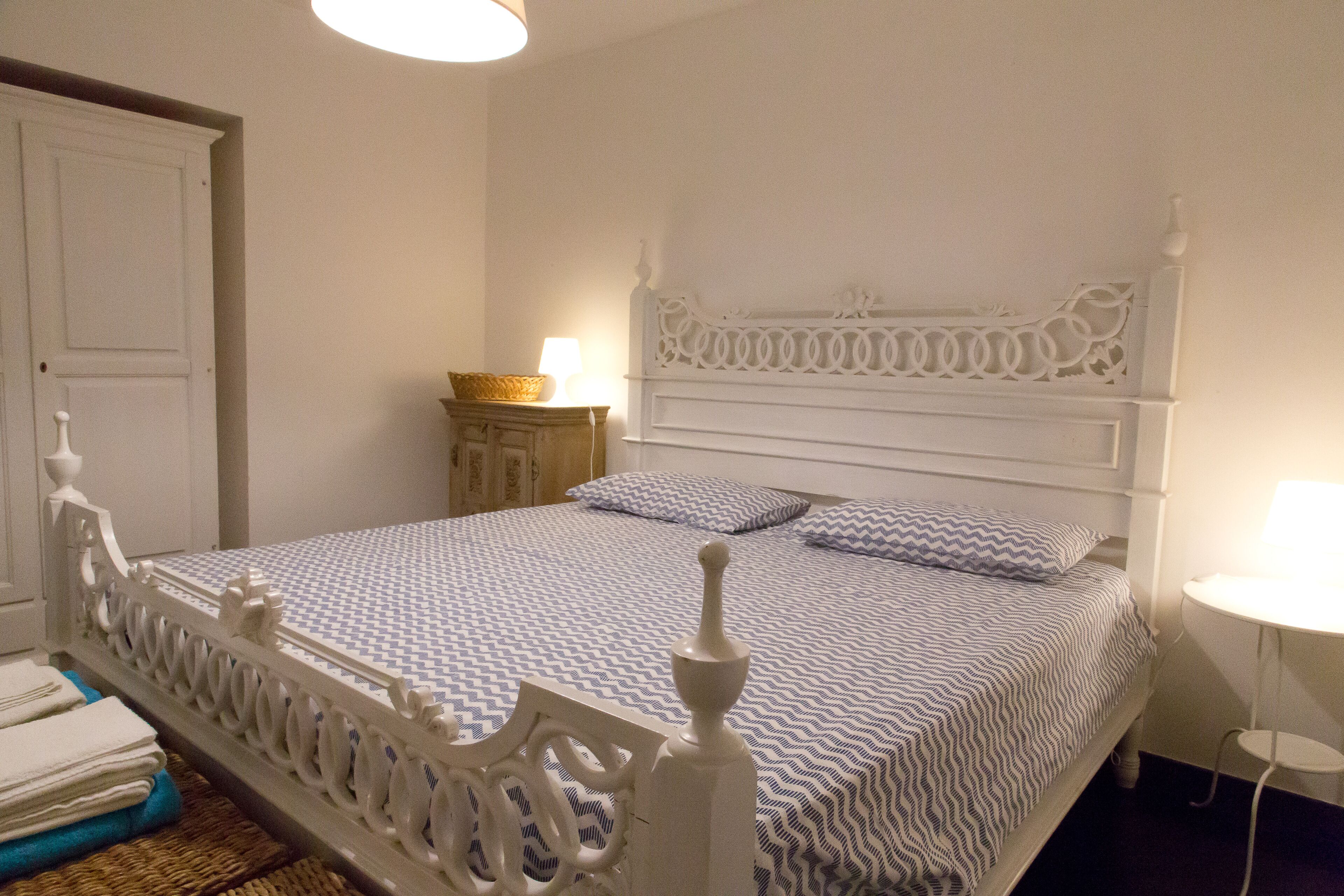 Una cama bien hecha con una colcha estampada, un cabecero blanco ornamentado y una mesita de noche con una lámpara.