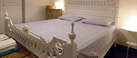 Una cama bien hecha con una colcha estampada, un cabecero blanco ornamentado y una mesita de noche con una lámpara.