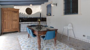A dining area with a wooden table and blue chairs, a kitchen in the background, and a window with a view.