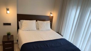 Ein ordentlich bezogenes Bett mit weißen Laken und einer dunkelblauen Decke, ein kleiner Nachttisch mit einer Pflanze sowie eine wandmontierte Leuchte.