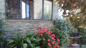 Un edificio de piedra con una ventana, una planta grande en maceta con flores rosadas y una pequeña mesa con una silla.