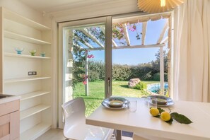 Comedor con una mesa dispuesta para dos, vista a un jardín a través de un ventanal, e interior blanco con estanterías.