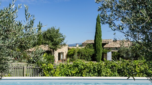 Une vue sur la piscine, avec un bâtiment en pierre, des oliviers et un ciel bleu azur.