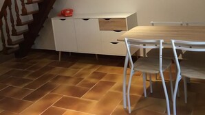 Un coin repas avec une table en bois et des chaises blanches, un meuble blanc avec des tiroirs, un objet rouge posé sur le dessus, et un escalier avec des balustrades en bois.