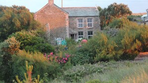 A stone house with a sloped roof, surrounded by dense vegetation and flowering plants.
