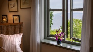 Een kamer met een rieten stoel, een kussen, een vaas met bloemen en een raam met uitzicht op water en groen.