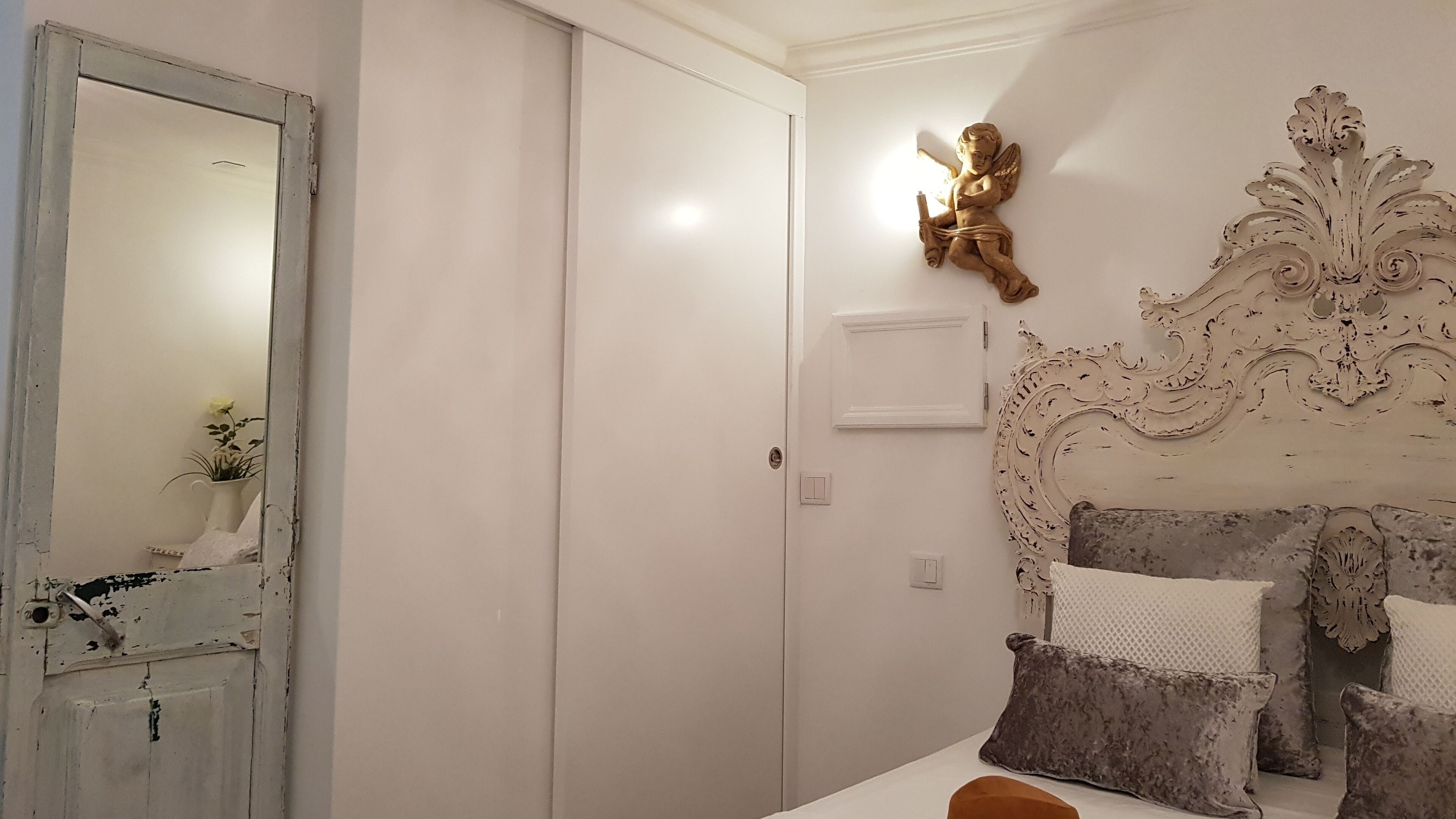 A bedroom with a white door, a decorative wall piece, a bed with multiple pillows, and a small table with a vase.