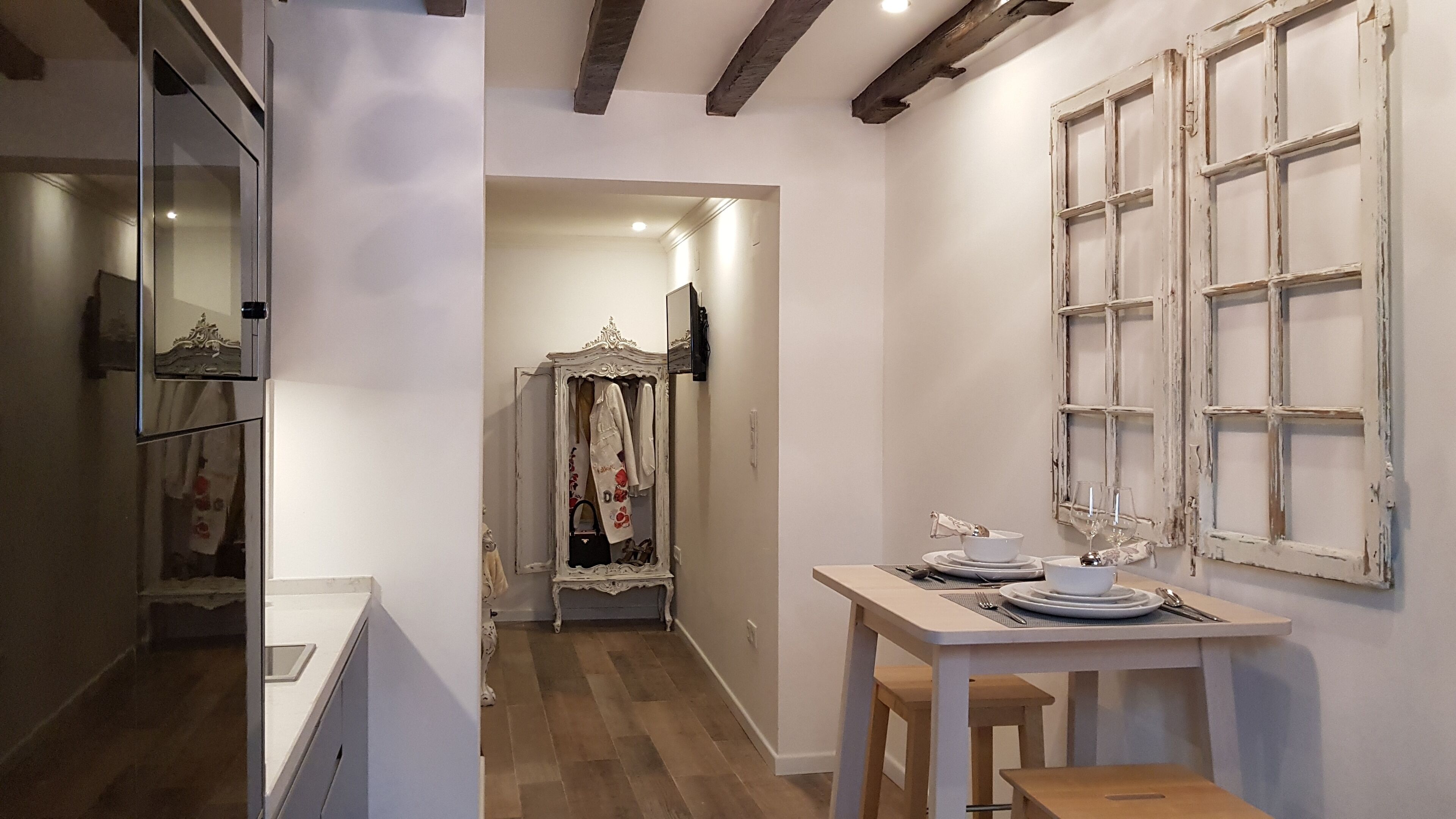 A narrow hallway with a wooden floor, a white door, and a mirror on the left. A table with plates and glasses is set up in the room.