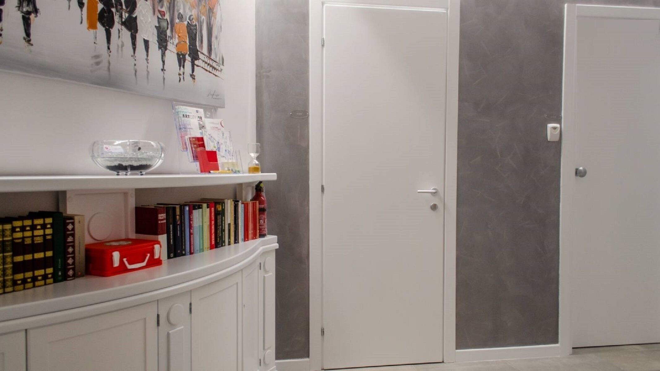 A room with a white door, a white cabinet with books, and a shelf with various items including a red box.