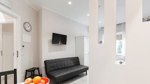 A modern living room with a sofa, a dining table with a bowl of fruit, a flat-screen TV mounted on the wall, and a window providing natural light. - Interior 1 Ciampino Rome with private balcony (CIAMPINO)