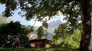 A rustic wooden cabin with a curved roof, surrounded by lush greenery and mountains in the background. - Beautiful 4* flat in a chalet with shared sauna, 300 meters from the slopes   (Les Avanchers-Valmorel)
