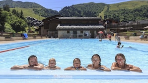 A swimming pool with clear blue water, surrounded by a wooden building with a sloped roof, set against a backdrop of green hills. - Beautiful 4* flat in a chalet with shared sauna, 300 meters from the slopes   (Les Avanchers-Valmorel)