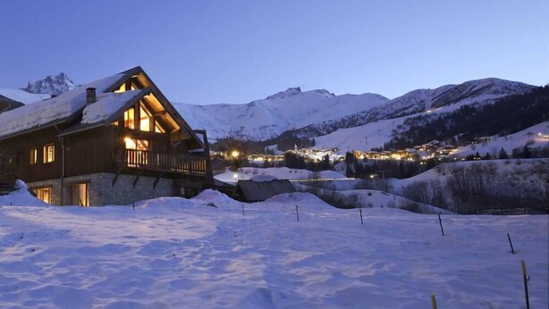 A wooden lodge with a balcony, surrounded by snow-covered mountains and a lit village in the background. - Beautiful 4* flat in a chalet with shared sauna, 300 meters from the slopes   (Les Avanchers-Valmorel)