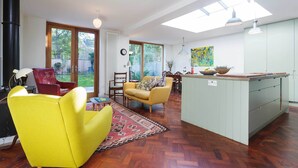 A modern living room with a yellow armchair, a red chair, and a sofa. There is a kitchen island with a sink and a dining area with a table and chairs. - East London Elegance, by Veeve (London)