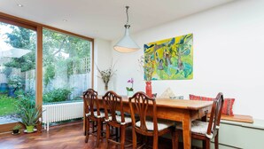 A dining area with a wooden table and chairs, a pendant light, a colorful abstract painting, and a view of a garden through large windows. - East London Elegance, by Veeve (London)