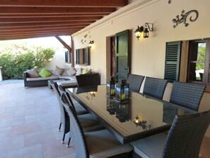 An outdoor patio with a glass-top table surrounded by wicker chairs, a sofa with cushions, and a door with green shutters.