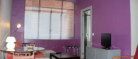 A living room with purple walls, a window with blinds, a television on a cabinet, a red chair, and a table with a lamp.