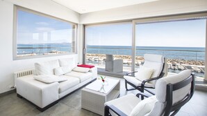 A modern living room with a white sofa, a white armchair, and a wicker coffee table. Large windows offer a view of the sea and a rocky shoreline. - Luxury house on the front line of the sea (Son Serra de Marina)
