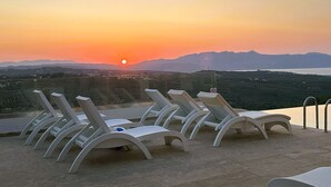 Una terraza con sillones blancos mirando una puesta de sol sobre un paisaje montañoso.