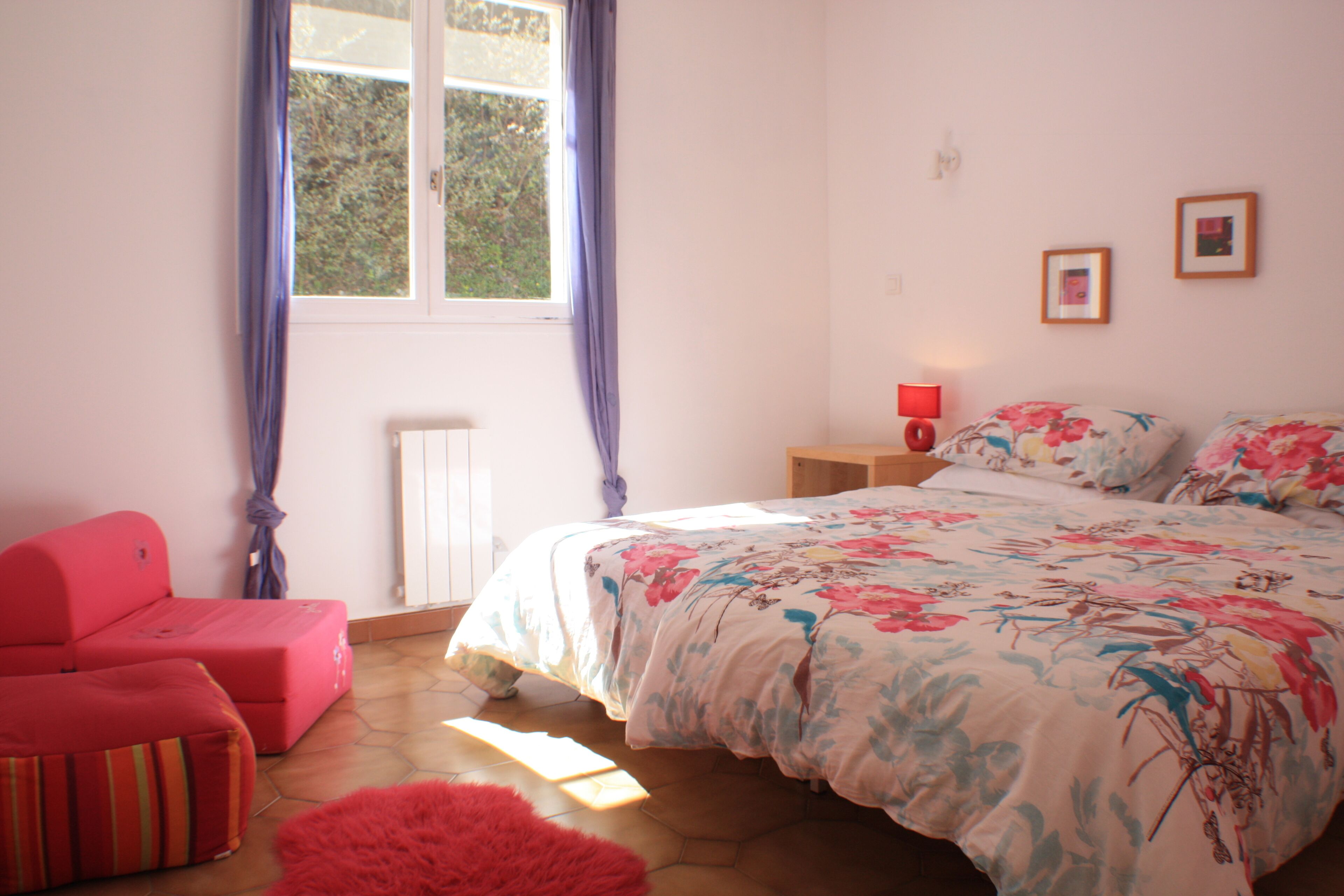 Une chambre à coucher avec un lit, une table de chevet, une lampe rouge, une chaise rose et une fenêtre avec des rideaux.