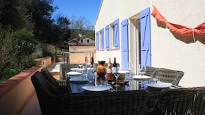 An outdoor dining area with wicker furniture, a table set with plates, glasses, and bottles, and a view of a building with blue shutters. - Large fully air conditioned luxury villa with pool in beautiful location (Reynès)