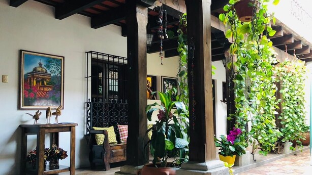 A covered outdoor area with a wooden bench, potted plants, and a wall-mounted painting. - Hotel La Casa de Don Moncho (Antigua Guatemala)