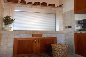 A kitchen with a stone backsplash, wooden cabinets, a window with a roller blind, and a coffee machine. - CASA SARADO (Polignano A Mare)