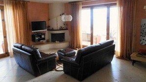 Una sala de estar con chimenea, sofás de cuero, televisor y ventanas grandes con cortinas.