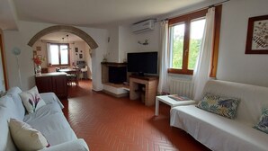 A living room with a sofa, a TV, and a fireplace. There is a kitchen in the background with a dining table and chairs.
