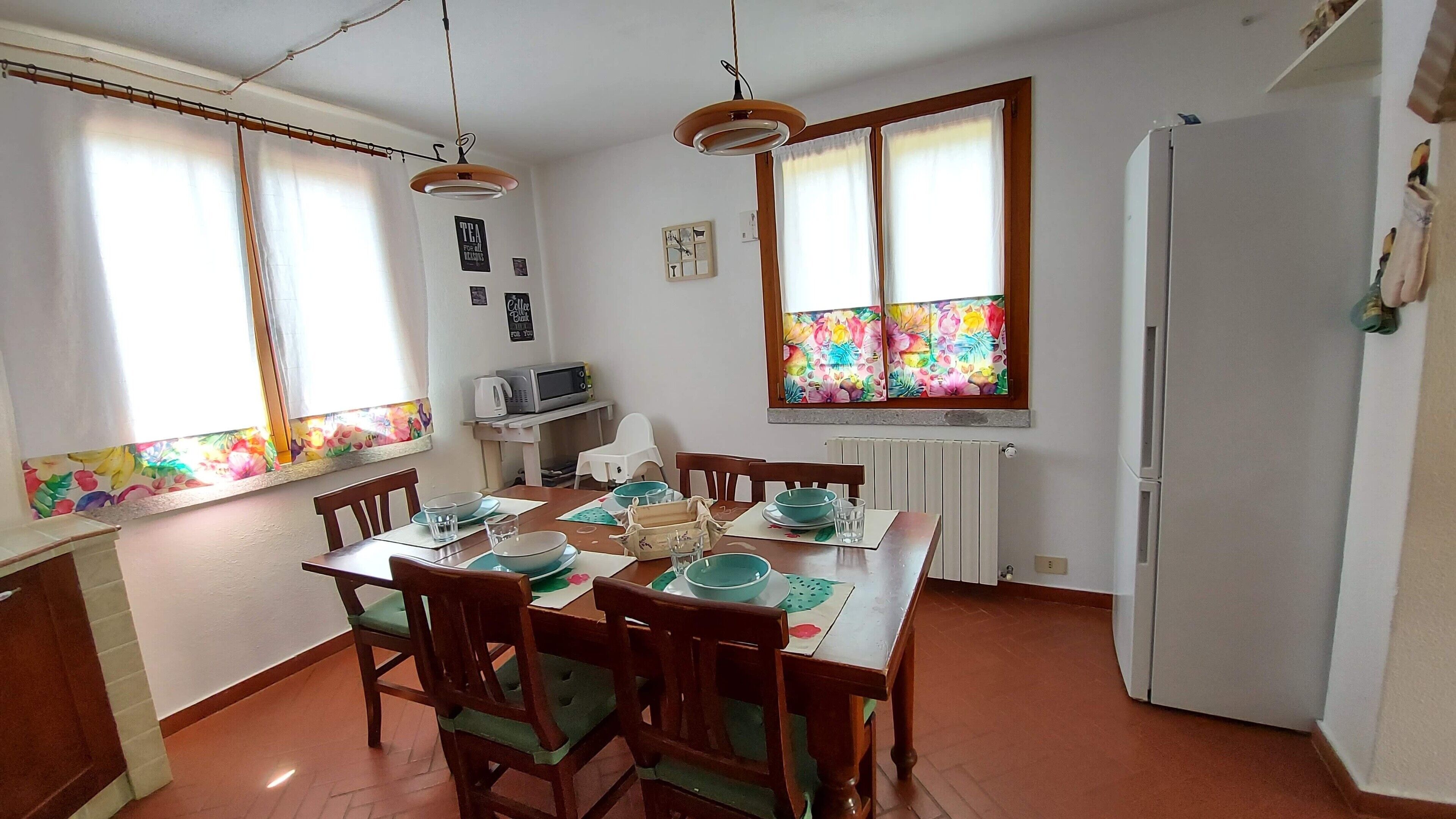 A dining area with a wooden table set for four, surrounded by chairs. There is a window with a colorful floral blind, a white refrigerator, and a microwave on a small table.