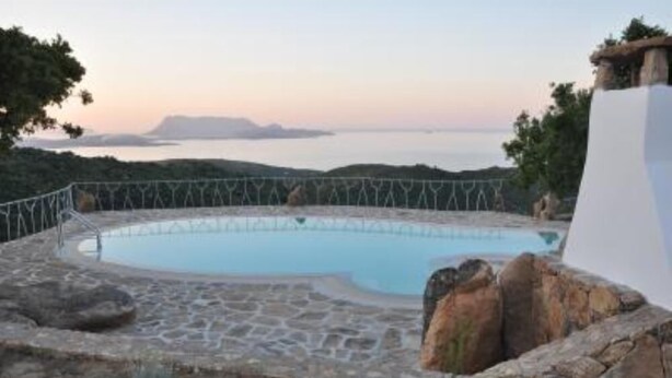 Una piscina de pedra amb tanca metàl·lica, envoltada de pedres i vegetació, amb vista a una massa d'aigua i terres llunyanes.