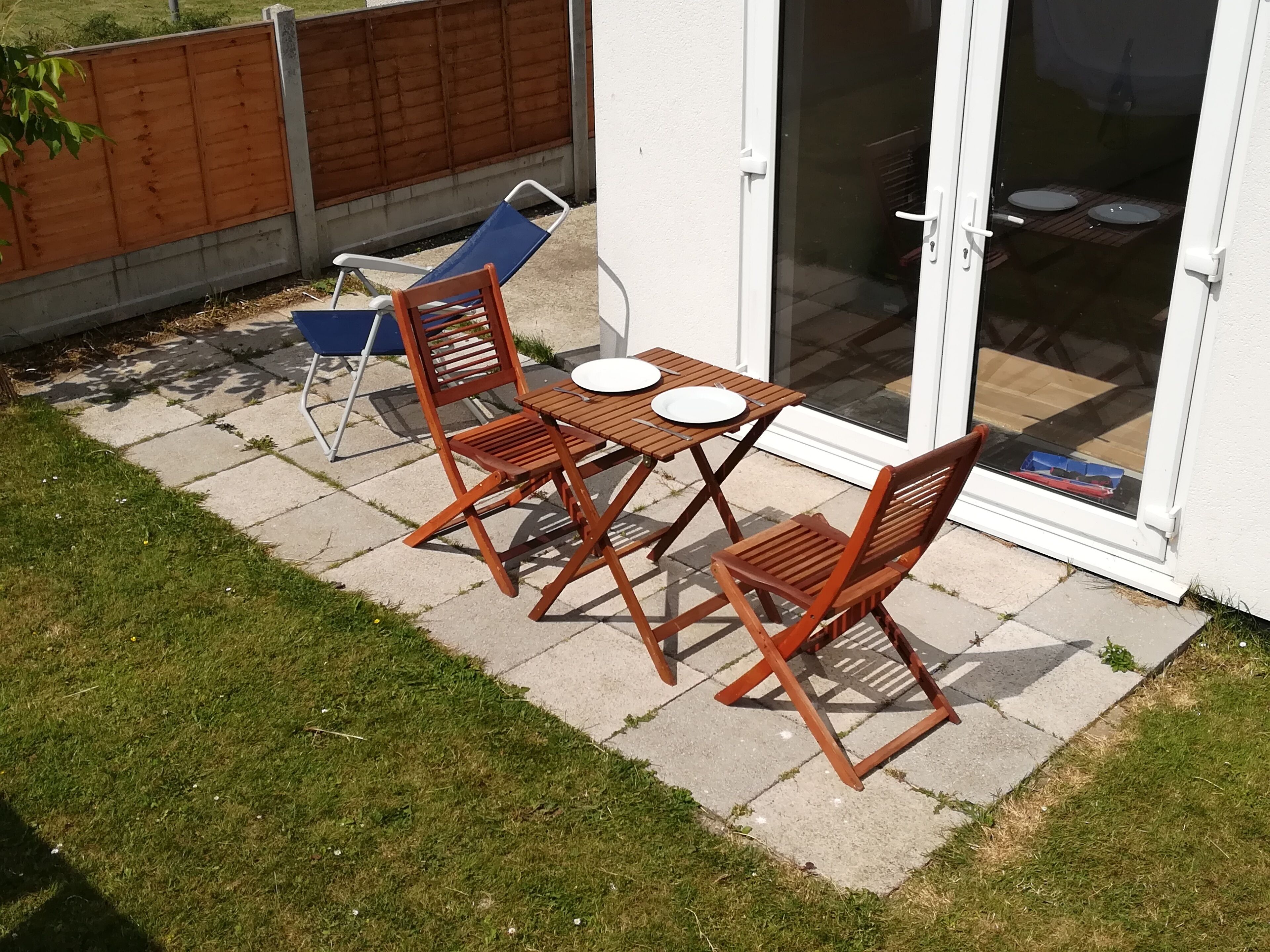 Un patio con una mesa y cuatro sillas de madera, una silla plegable azul y un cerramiento de vidrio corredero que da a una zona cubierta con dos platos blancos en el suelo.