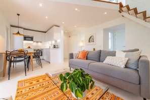 Ein modernes Wohnzimmer mit grauem Sofa, einem Essbereich mit Stühlen, einer Küche im Hintergrund und einer Treppe, die in ein Obergeschoss führt.