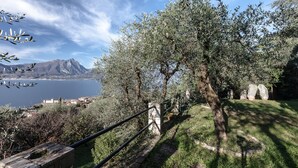A view of a coastal area with olive trees, a stone pathway, and a distant view of a castle by the sea. - Villa I Gelsomini,  6 Bedrooms, Garden, Terrace, Stunning View of Lake Garda (TORRI DEL BENACO - VENETO)